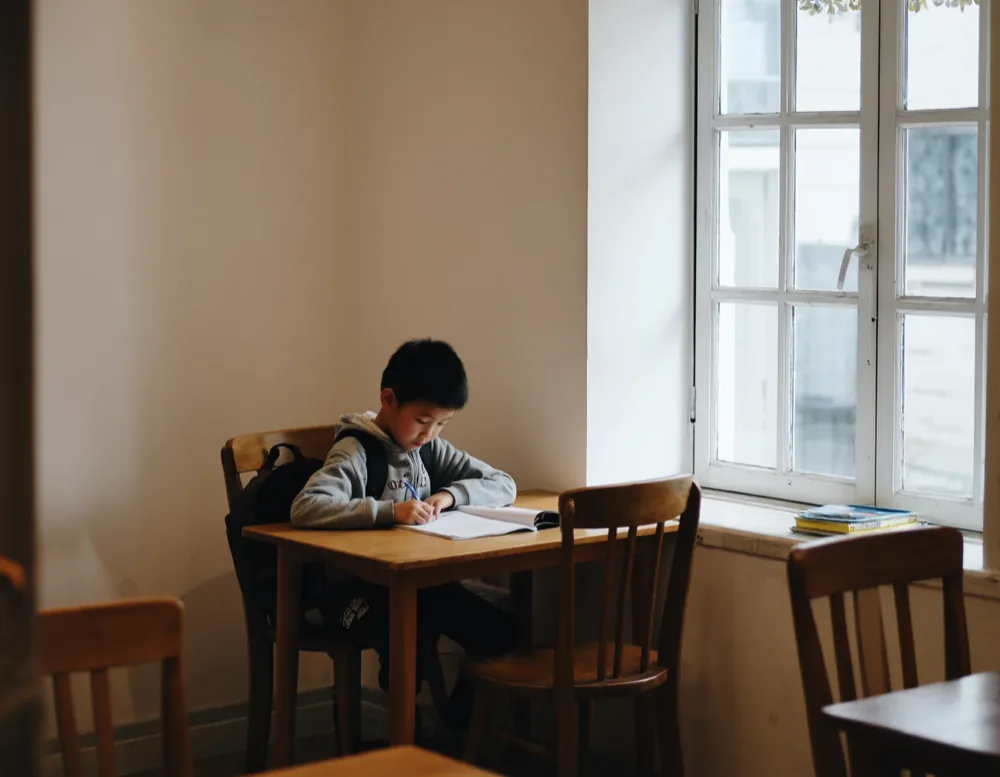 A boy in a grey hoodie doing his homework alone at a small table by the tall window