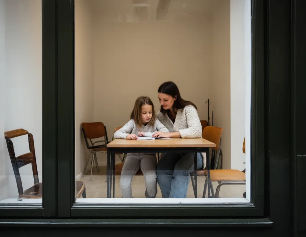 Seen through the shop window: a mother and her daughter sitting side by side at one of the tables, looking at an exercise book together